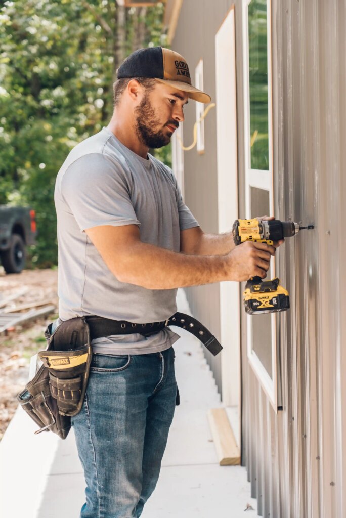 A man wearing a cap and tool belt uses a yellow power drill to install screws into the metal siding of a building. Trees and construction materials are visible in the background.