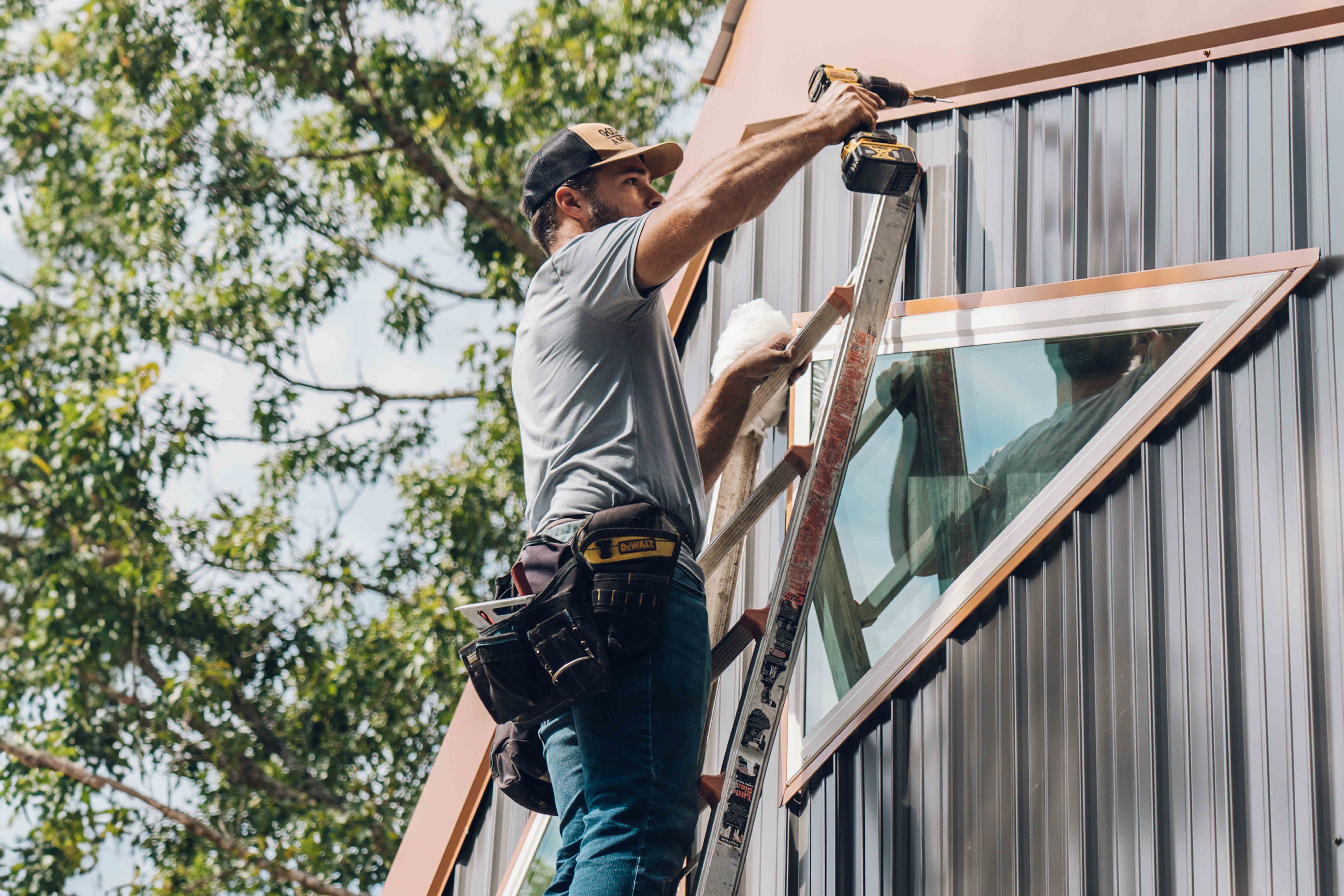 A construction worker stands on a ladder, installing or fixing a window on a modern building with metal siding, using a power drill. Trees with green leaves are visible in the background.