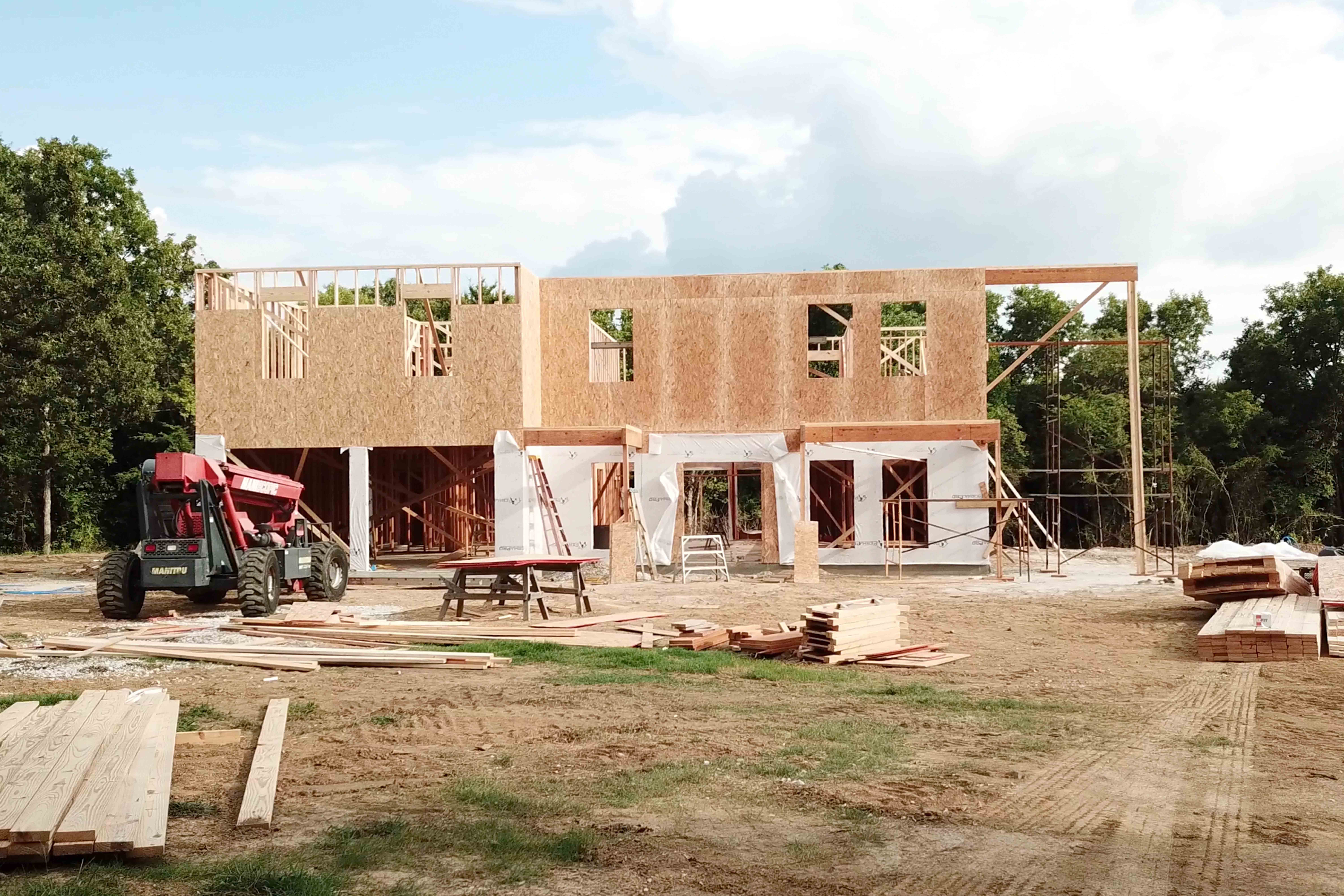 A two-story house under construction with exposed wooden framing, scaffolding, and building materials scattered around. A red forklift is parked nearby, and trees are visible in the background.